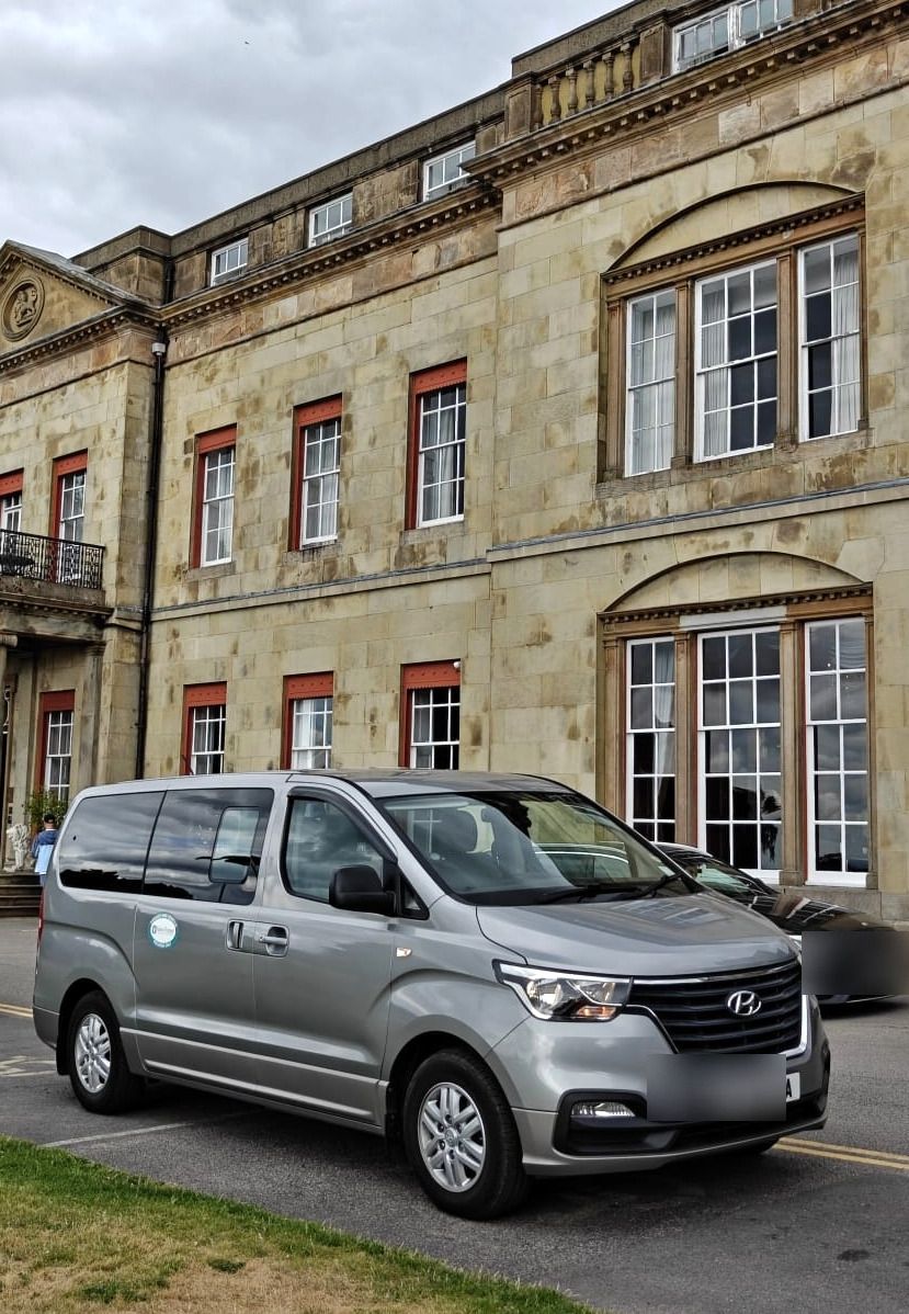 A silver minibus arriving at the front of a large country house.