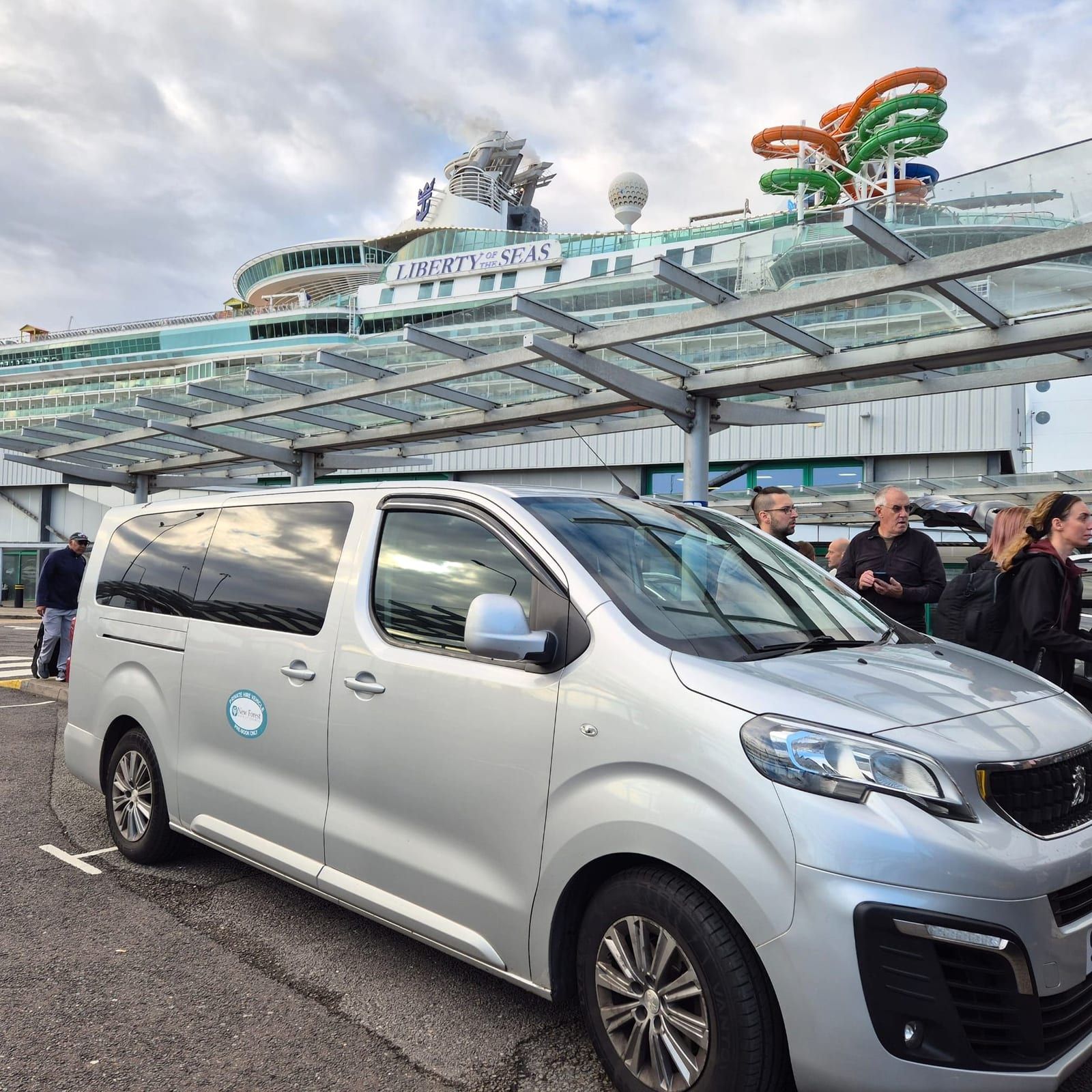 A silver minibus by a cruise terminal canopy with a docked cruise ship behind.