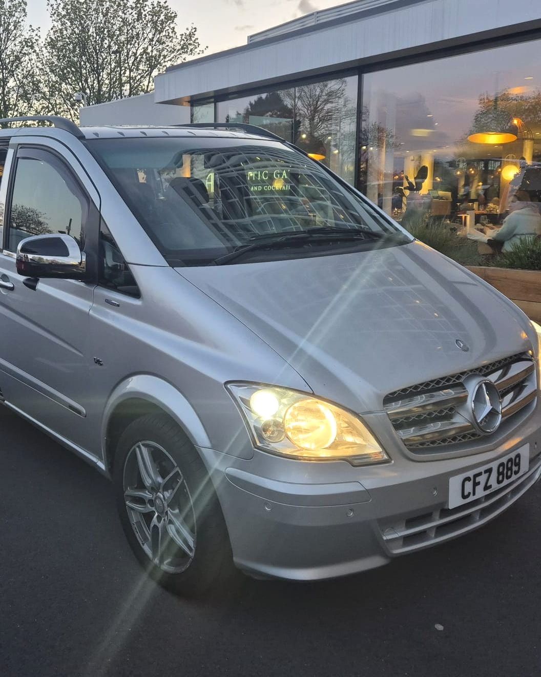 A silver minibus with its headlights on at dusk.