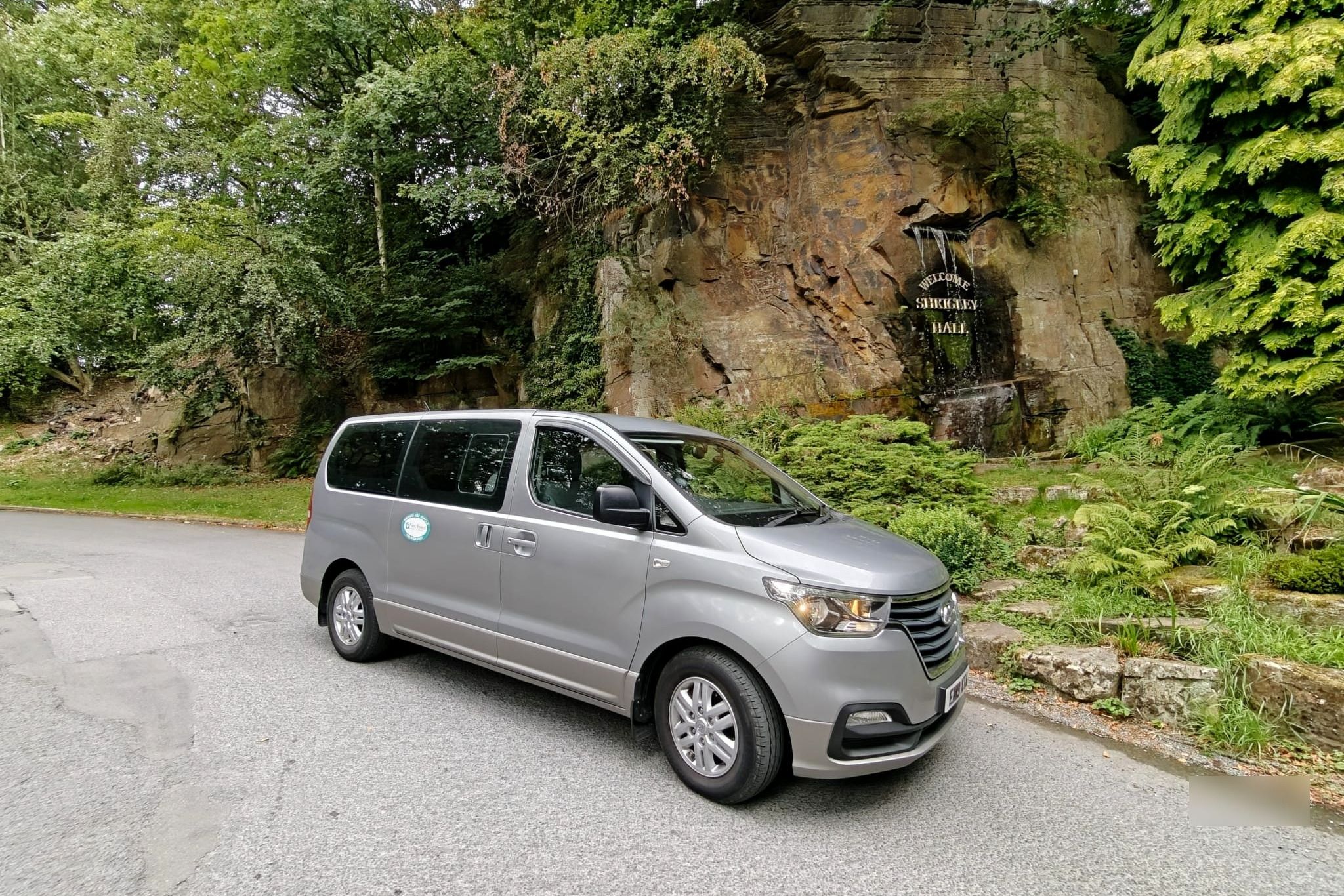 A silver minibus parked on a driveway beside a landscaped rock garden.
